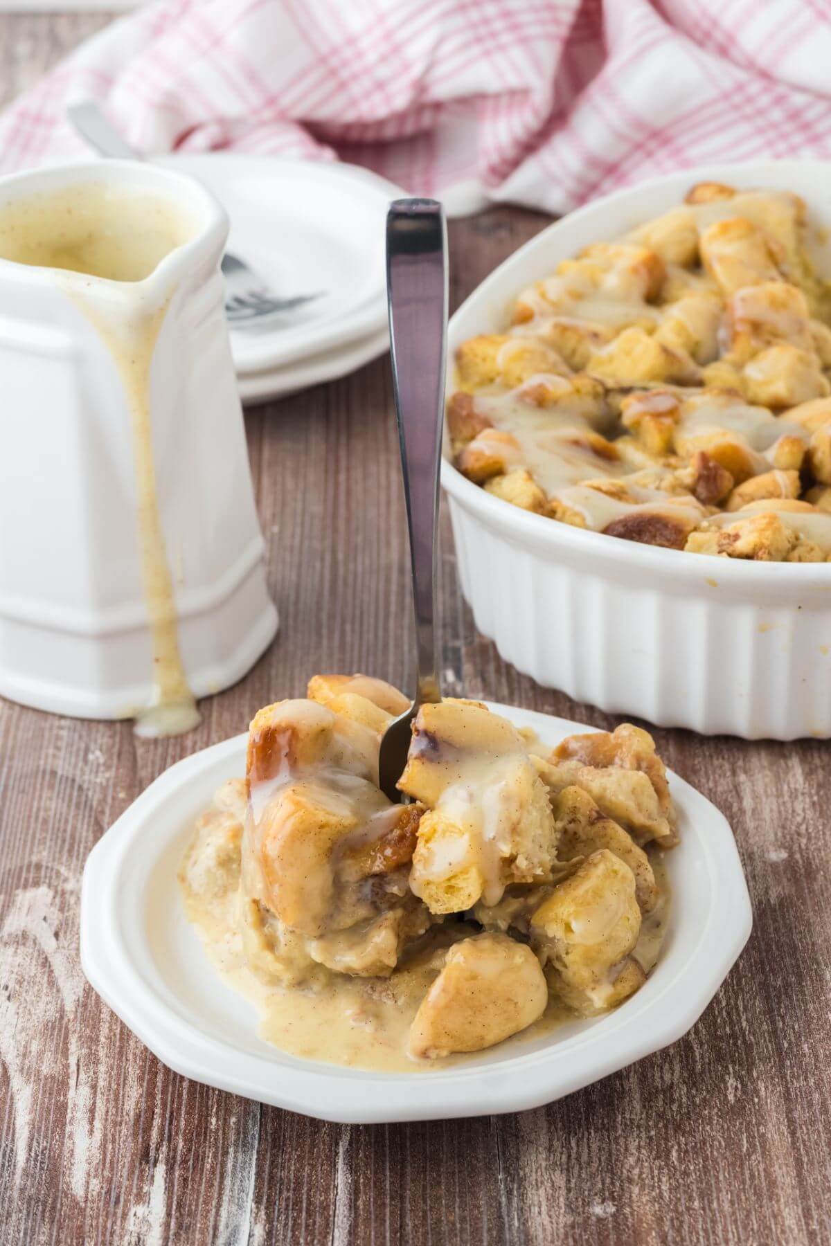 A white pitcher of glaze, white baking dish full of pudding, and a white plate filled with cinnamon roll chunks with a fork sticking up straight are all on a wooden table.