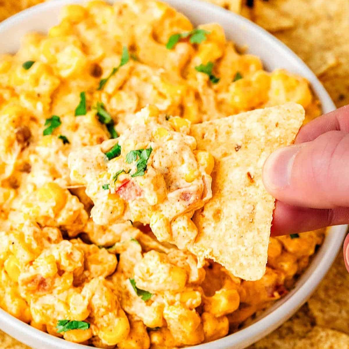 Three cheese hot corn dip in a bowl with a hand holding a chip dipping.
