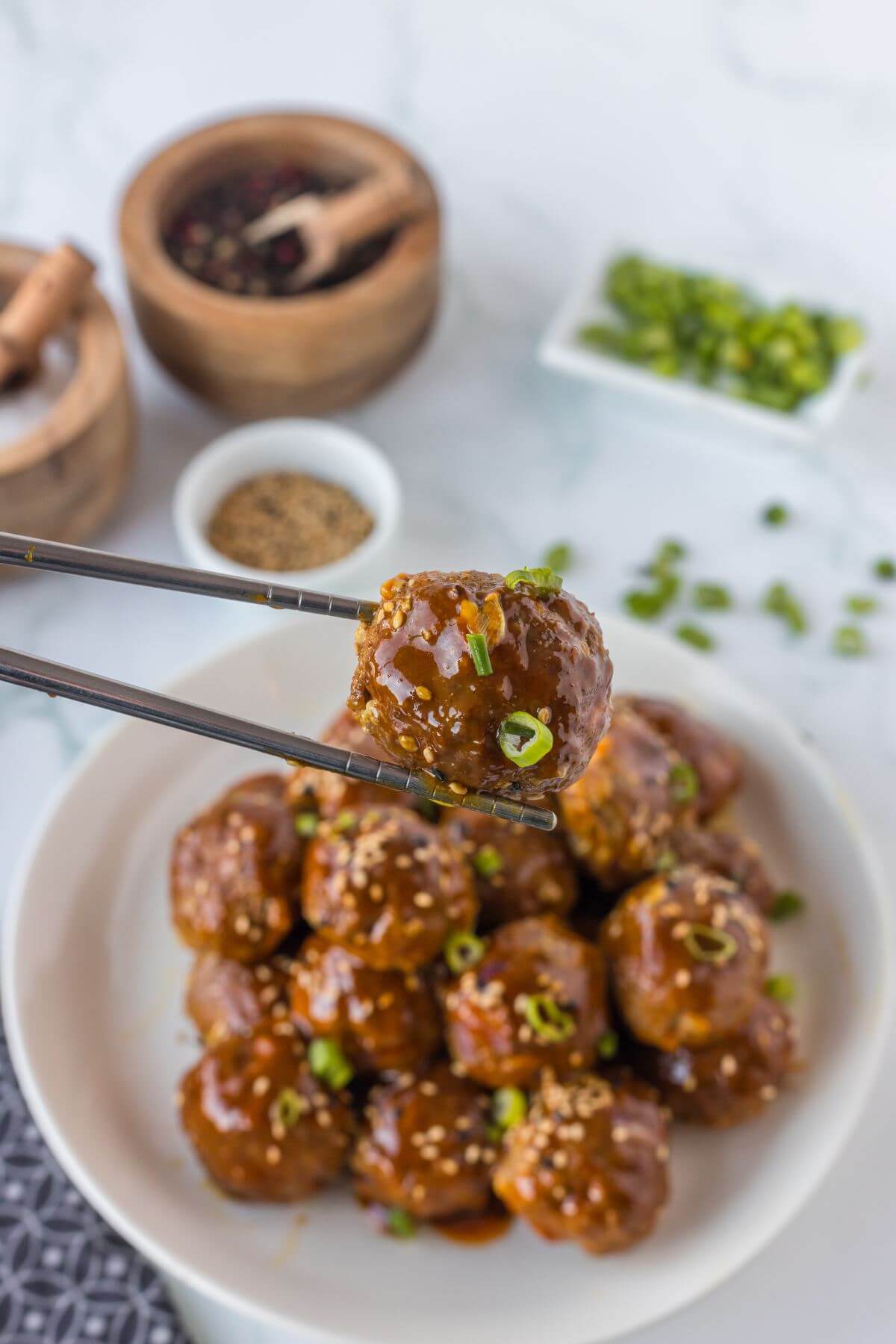 Chopsticks hold up a meatball at an angle to better show green onion and sesame seed garnish above a full dish.