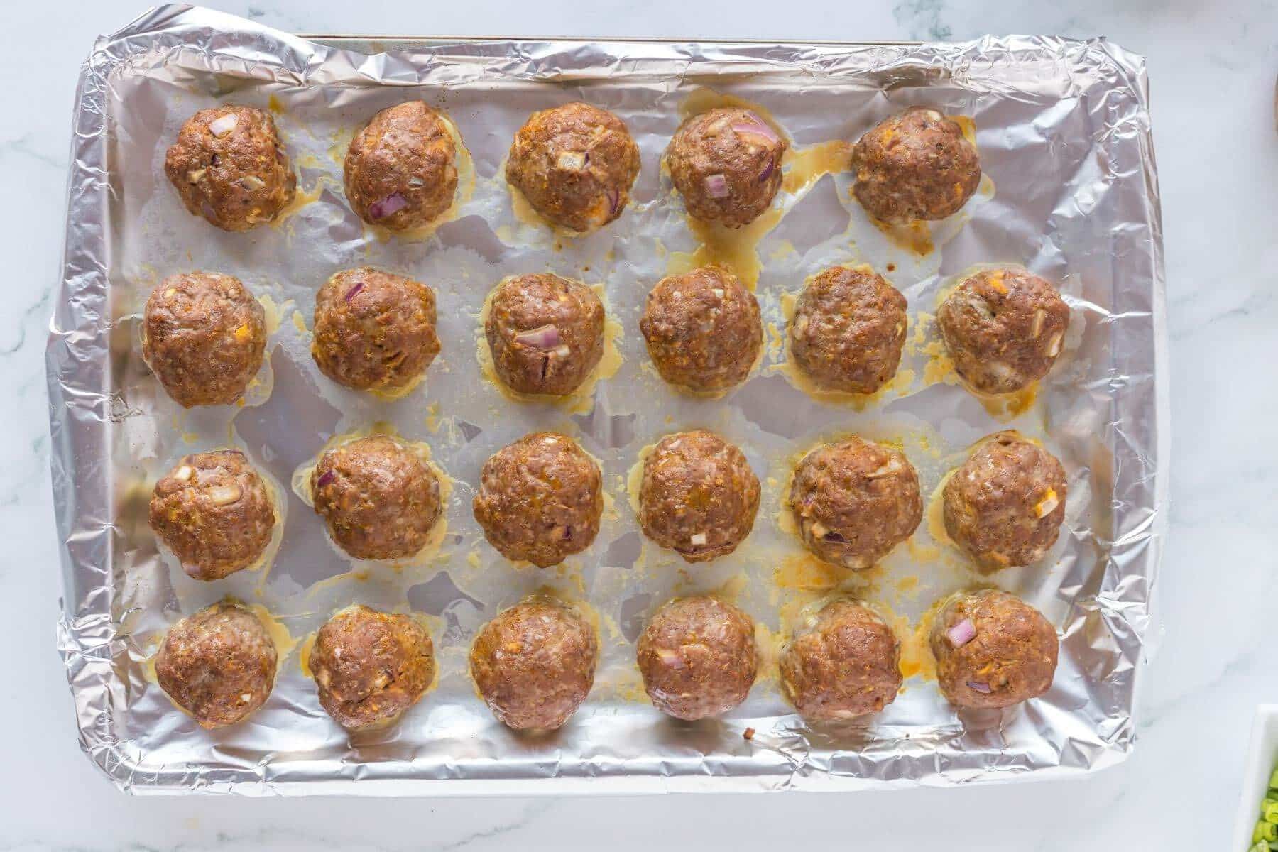 Cooked meatballs lay in neat rows on a pan lined with aluminum foil.