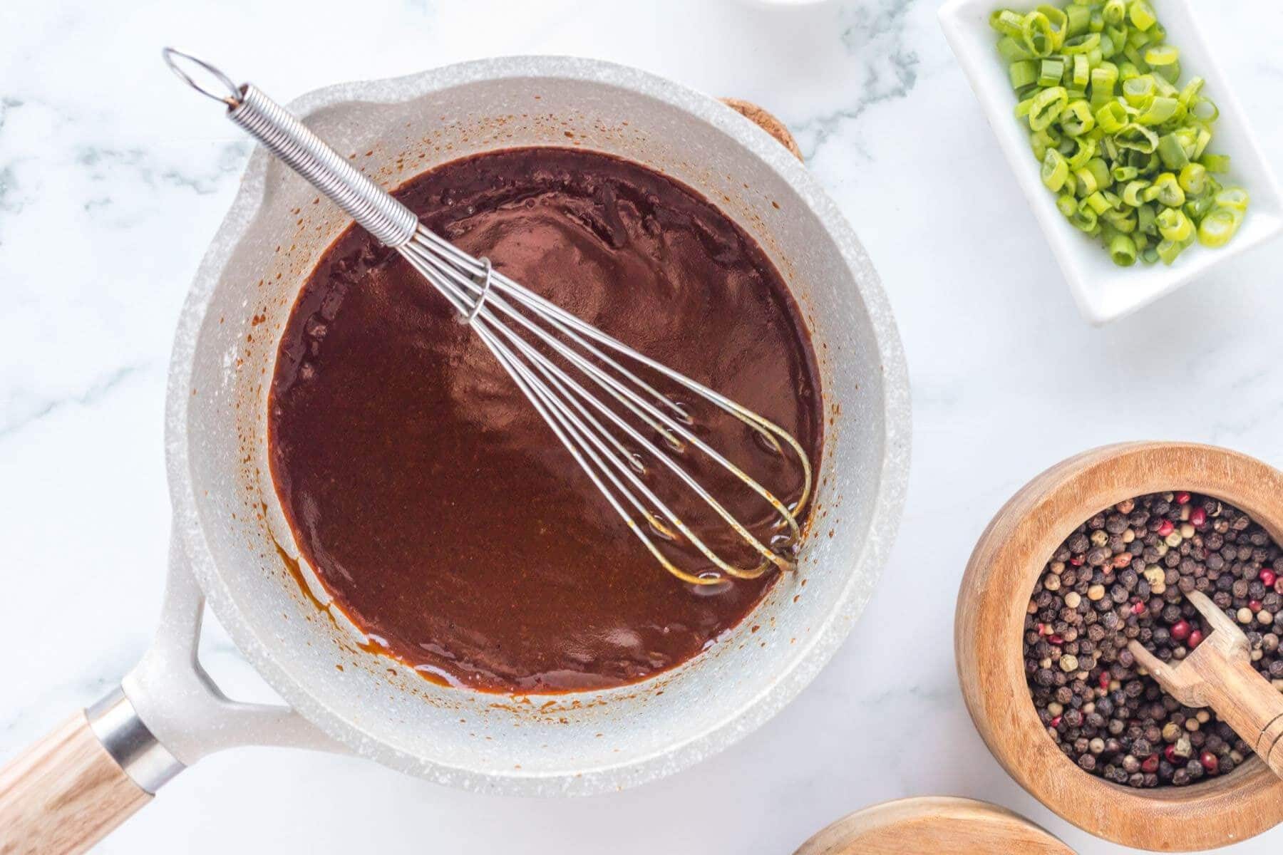 Thick brown glaze sits in a saucepan with a wire whisk on top.