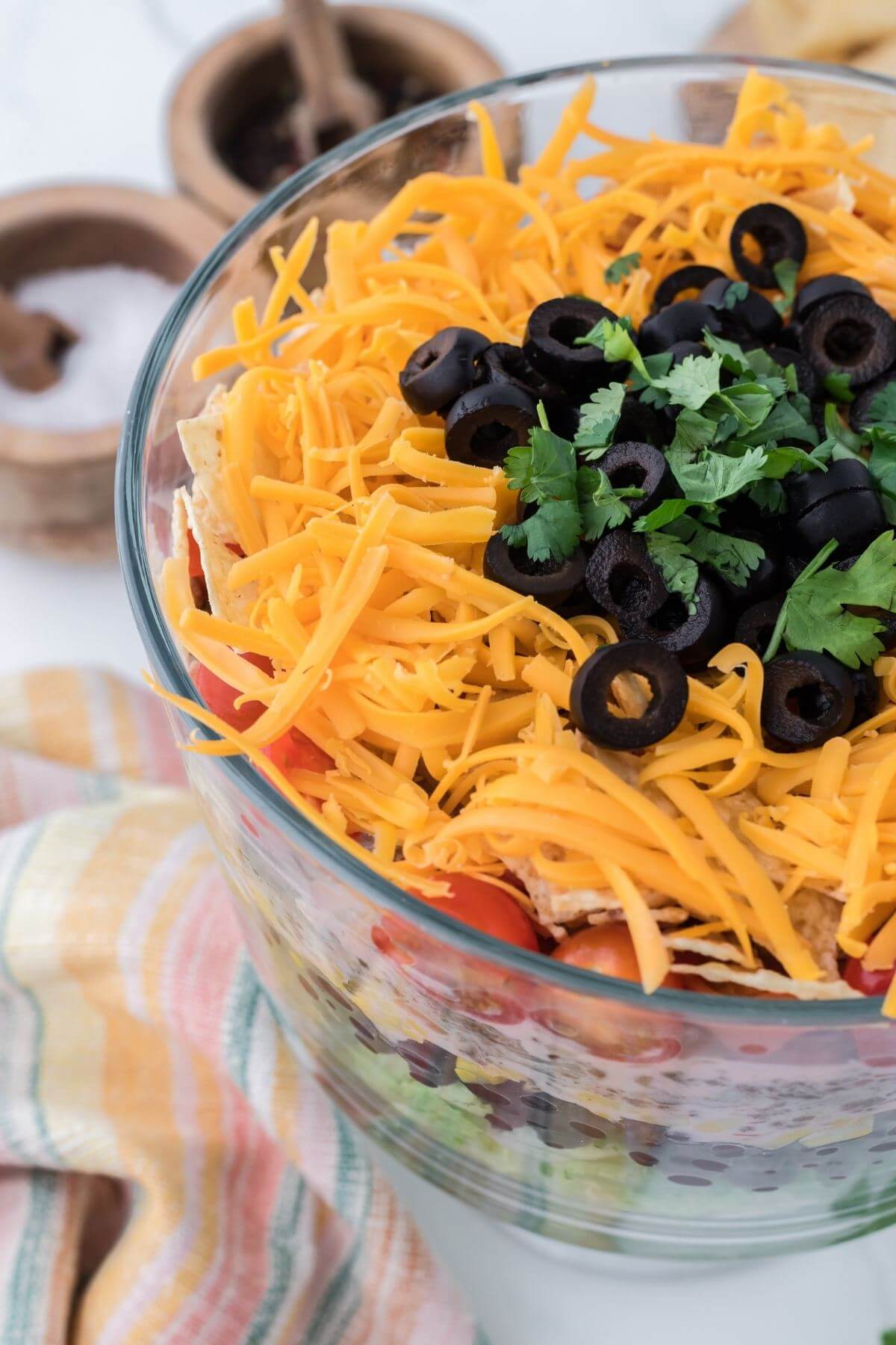 A colorful striped cloth sits next to a deep glass bowl of salad layers topped with shredded cheese.