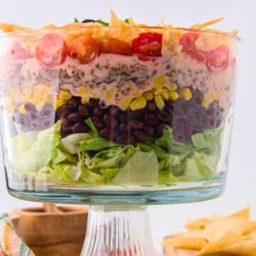 Layered taco salad in a pretty glass footed bowl with tortilla chips behind, for Facebook.