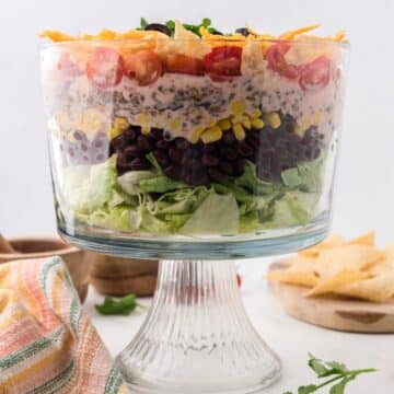 Layered taco salad side view in glass, footed bowl with tortilla chips nearby.