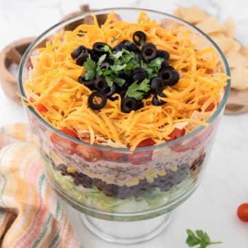 Layered Taco Salad in a glass footed bowl with tortilla chips in the background.