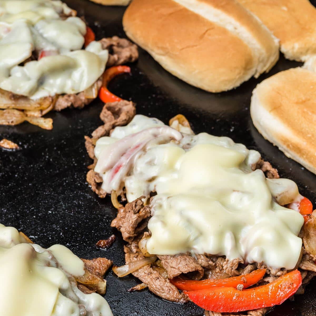 White cheese melting onto piles of Philly Cheesesteak mixture with rolls toasting in background. 