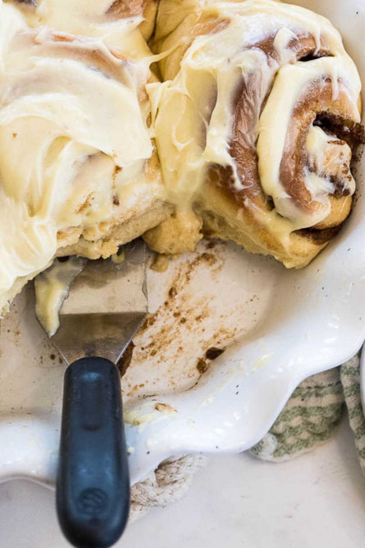 A metal spatula hangs out from under the bottom of cinnamon buns in a white decorative bowl.