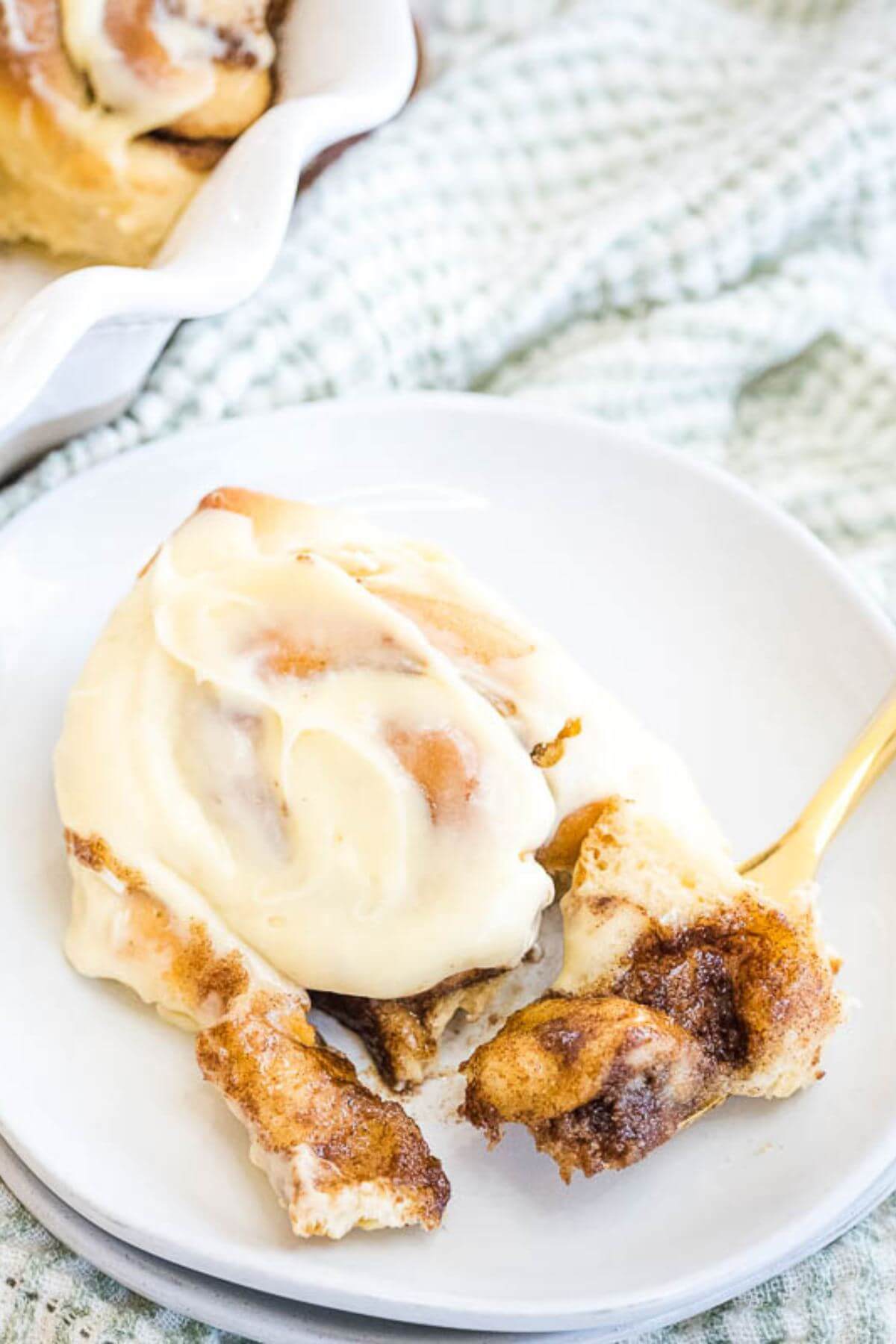 A golden fork lifts up a gooey chunk of roll covered in cinnamon next to the whole iced pastry.