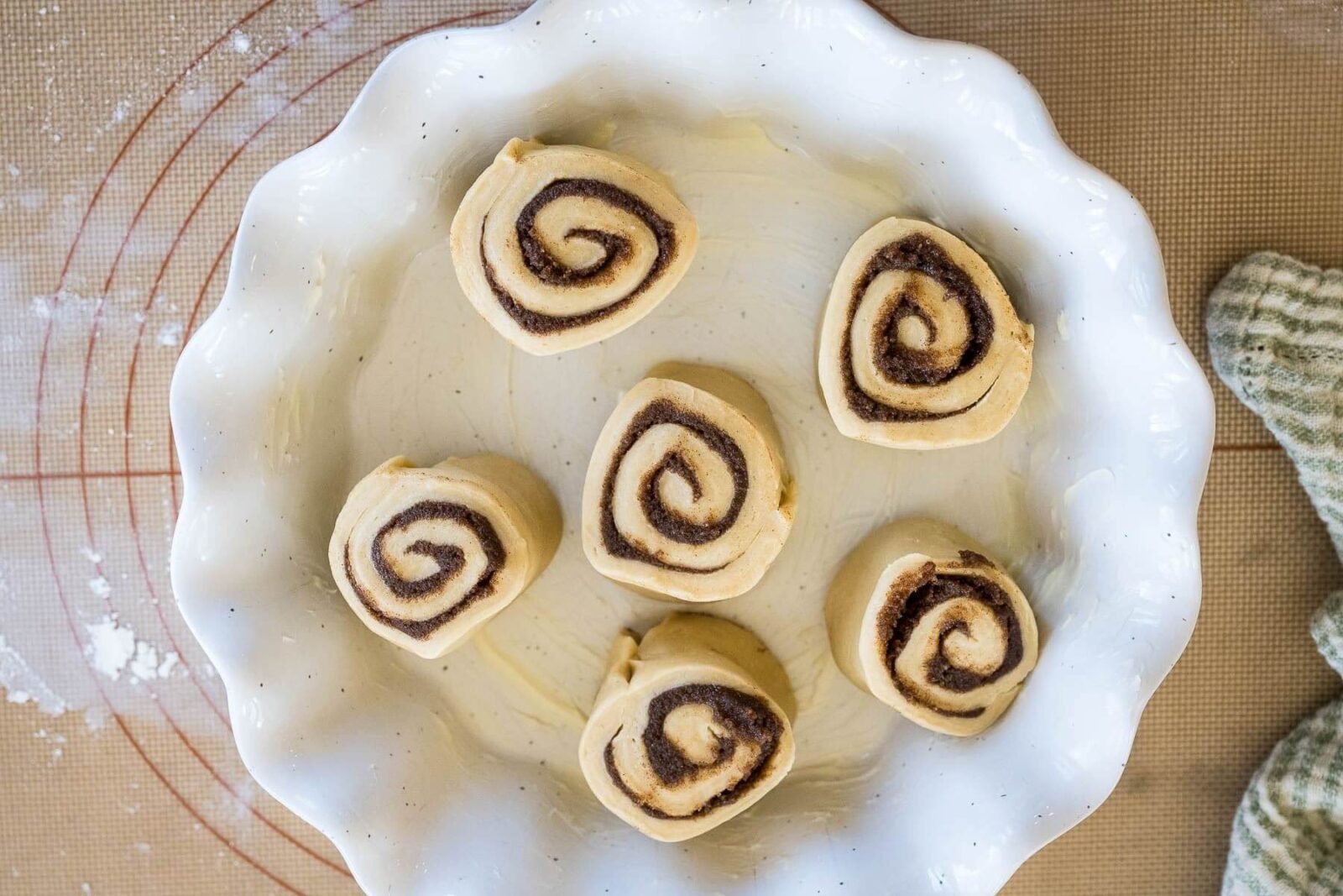 Unbaked, unrisen rolls with dark cinnamon swirls are placed a few inches from each other in a white wavy serving dish.