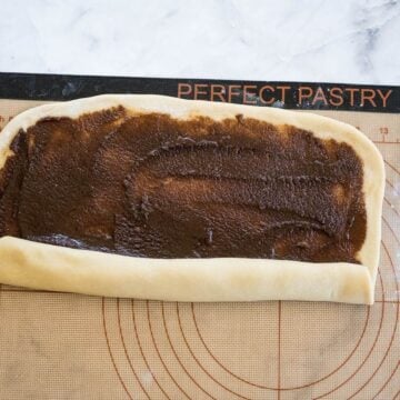 A slab of cinnamon covered dough has one long side starting to roll up.