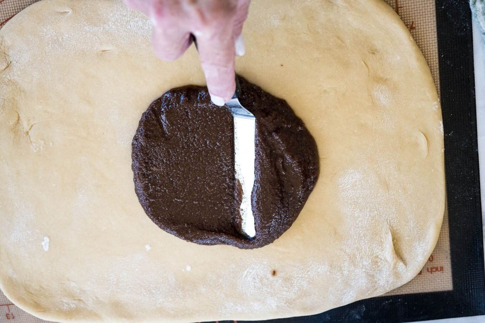 A hand uses a thin metal spatula to start spreading out dark brown cinnamon filling onto dough.