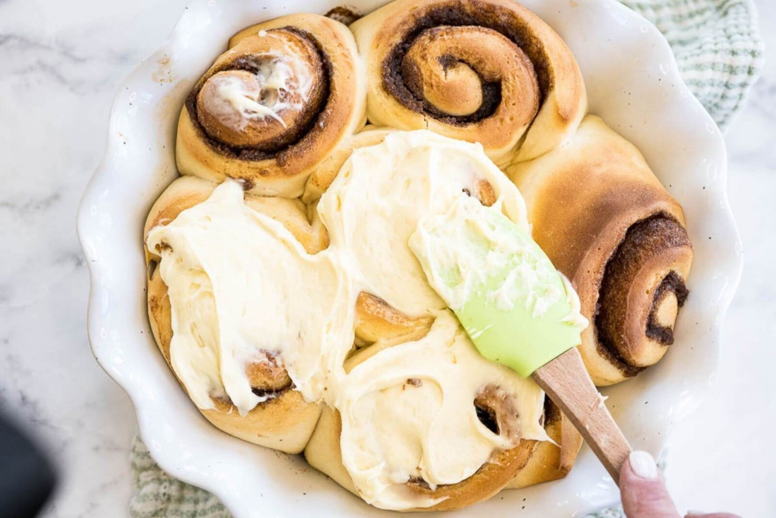 A hand uses a spatula to spread icing on top of cinnamon buns in a white dish.