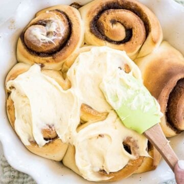 A hand uses a spatula to spread icing on top of cinnamon buns in a white dish.