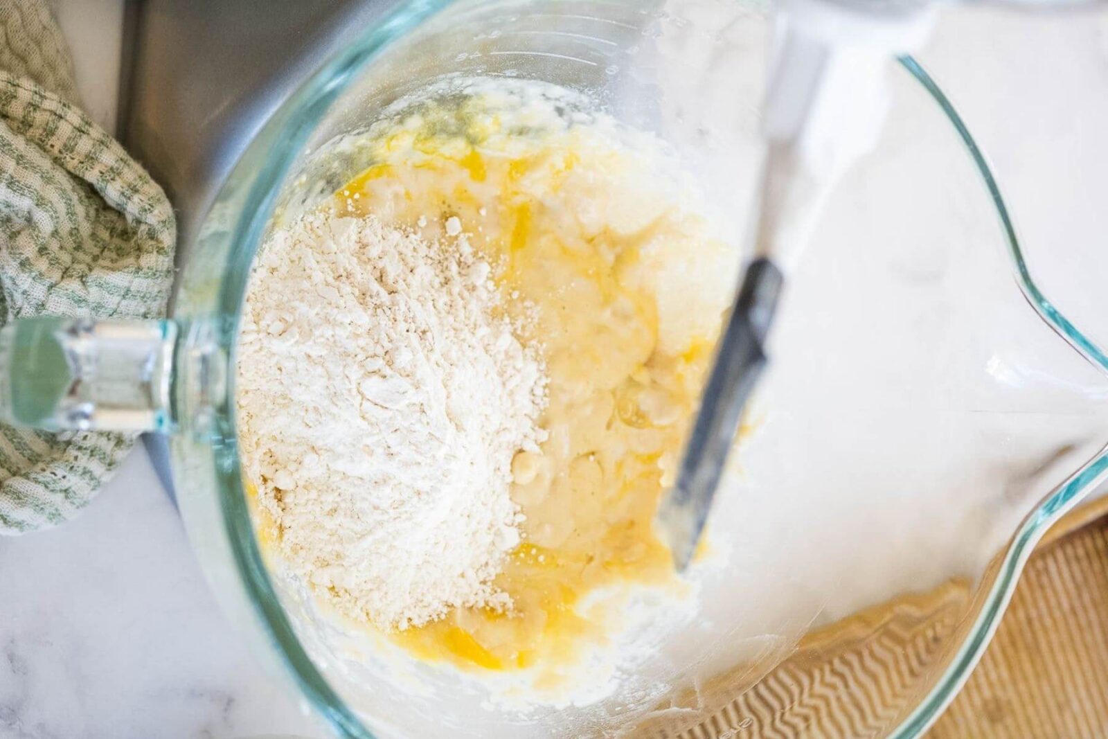 Flour sits on top of mixed dough ingredients in a glass mixing bowl.