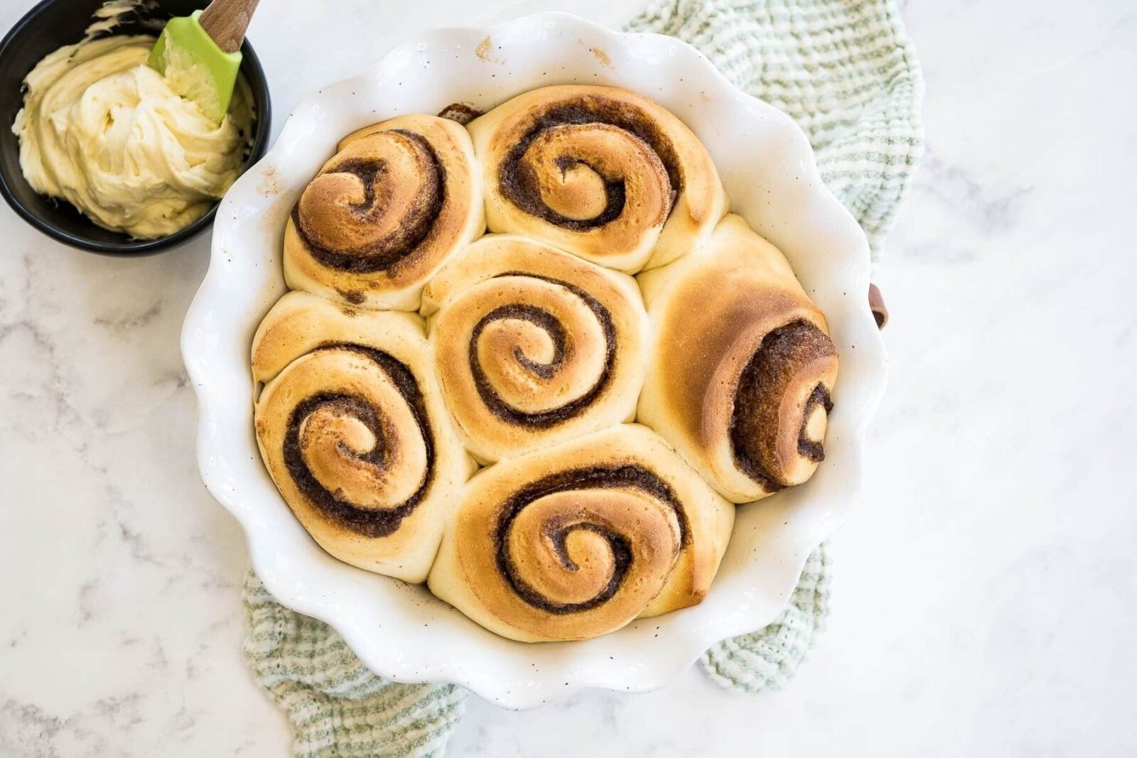 A white decorative serving bowl is filled with golden baked rolls with cinnamon swirl next to icing bowl.
