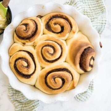 A white decorative serving bowl is filled with golden baked rolls with cinnamon swirl next to icing bowl.
