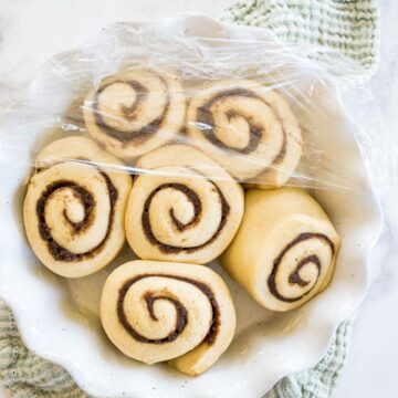 Unbaked rolls with cinnamon swirls sit together in a white baking dish under plastic wrap.