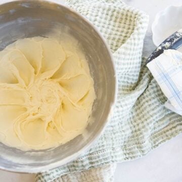 A metal bowl from a stand mixer is filled with cream cheese icing next to a dish cloth and spatula.