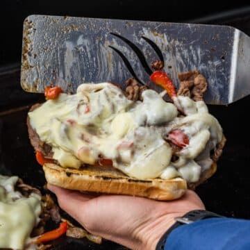 Hand holding a Blackstone spatula scooping Philly Cheese Steak filling onto a toasted bun.