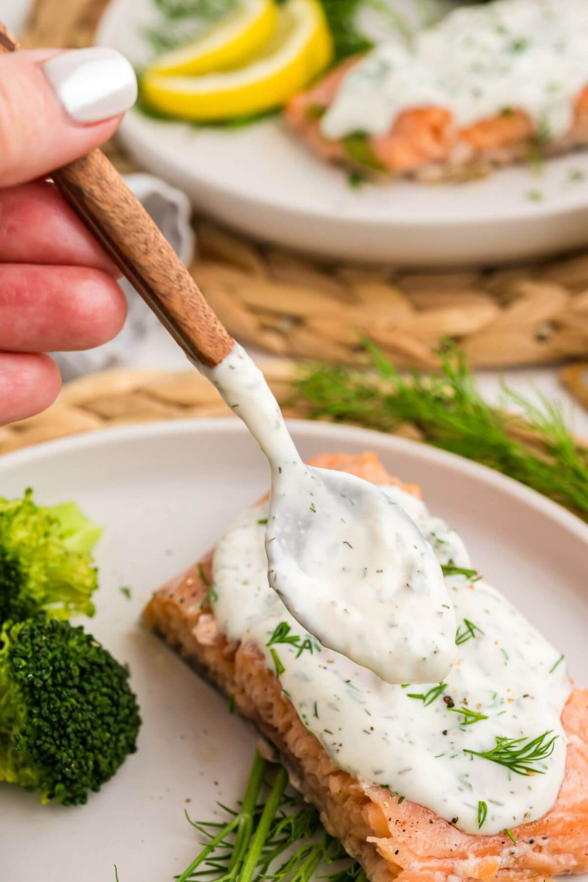 A hand uses a spoon to add white lemon dill dressing to the top of a fish filet.