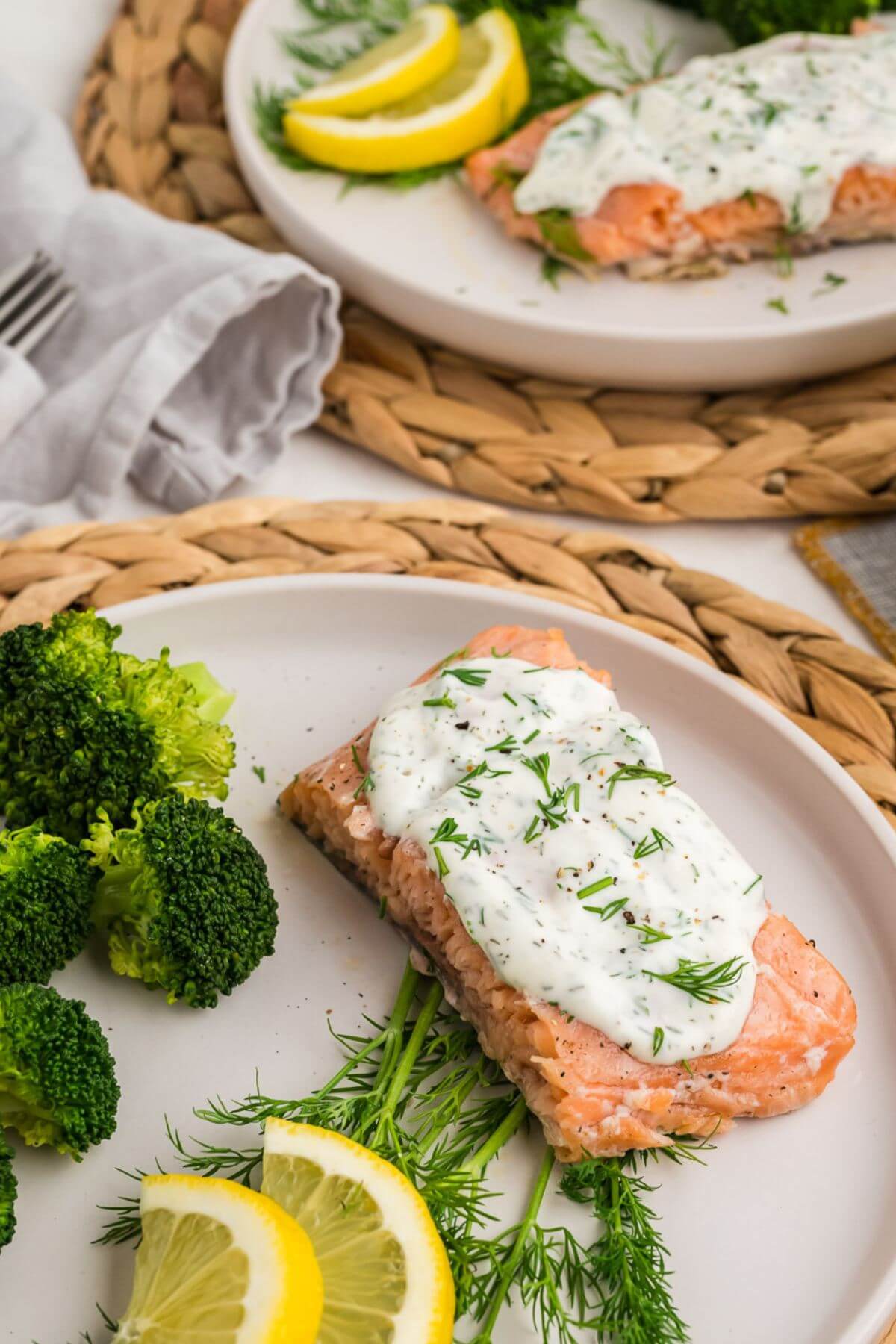 Two plates are filled with meat covered in a white sauce with green herbs all next to lemon slices and dill.