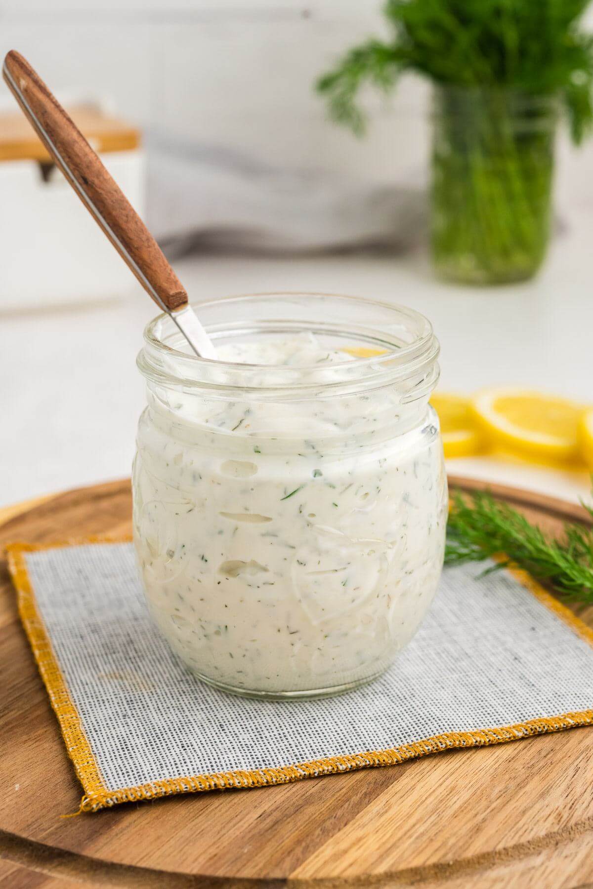 A jar of creamy white sauce with dill flecks is centered on a round wooden board.