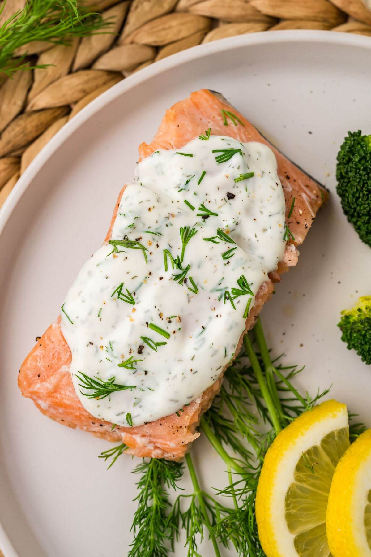 A salmon filet is coated with a white sauce with fresh dill sprinkled on top next to more dill and lemons.
