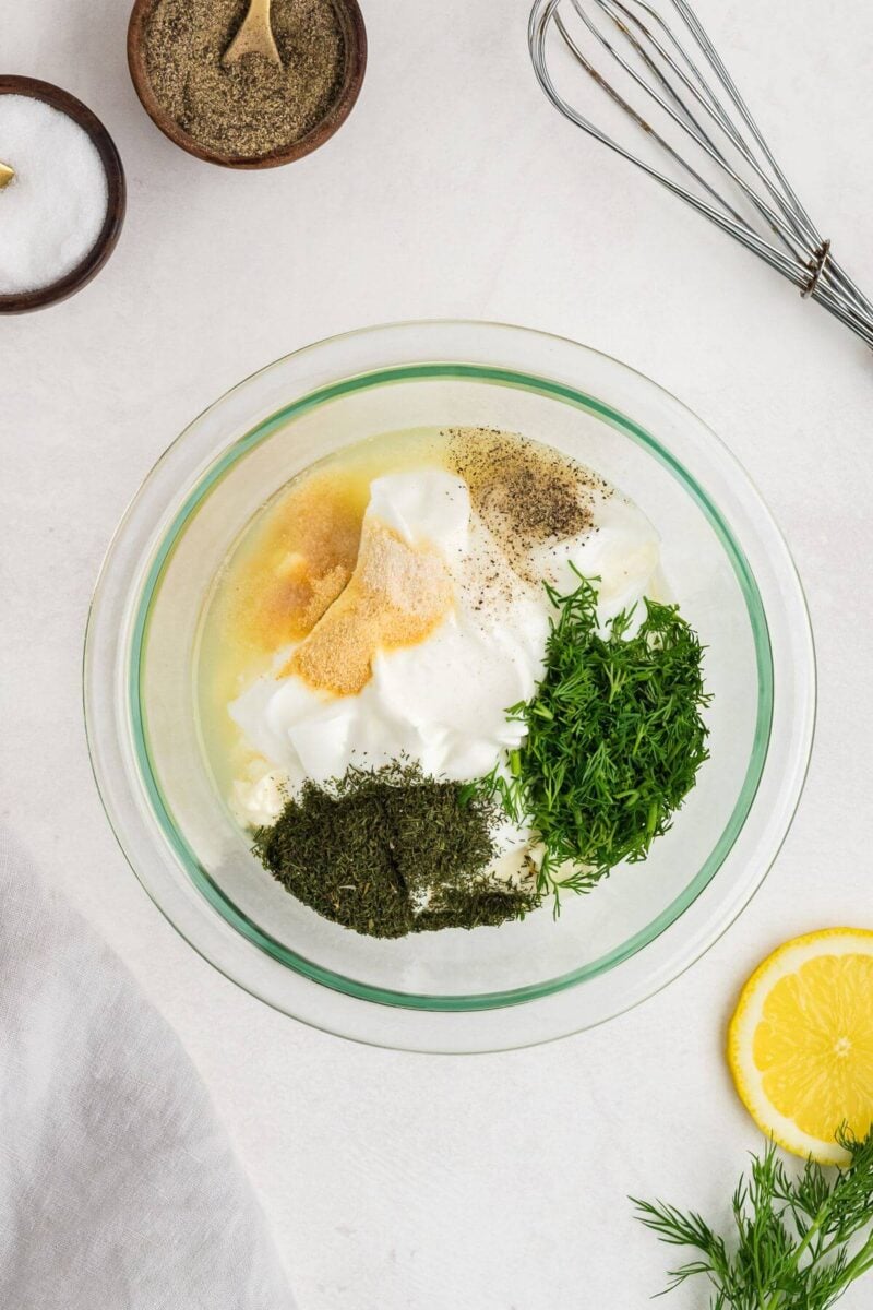 A mixing bowl is filled with a thick white base, green fresh and dried dill, and yellow and black spices on top.