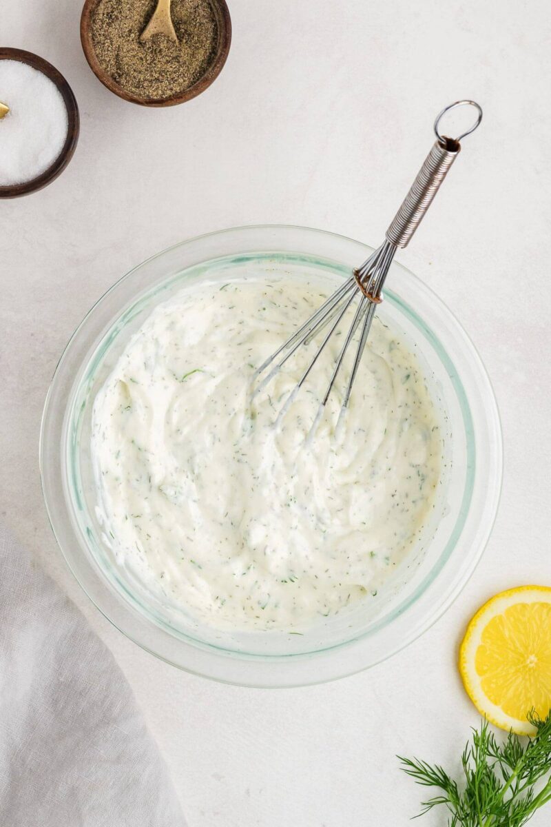 A whisk rests in a thick white sauce with green herb specks in a mixing bowl.