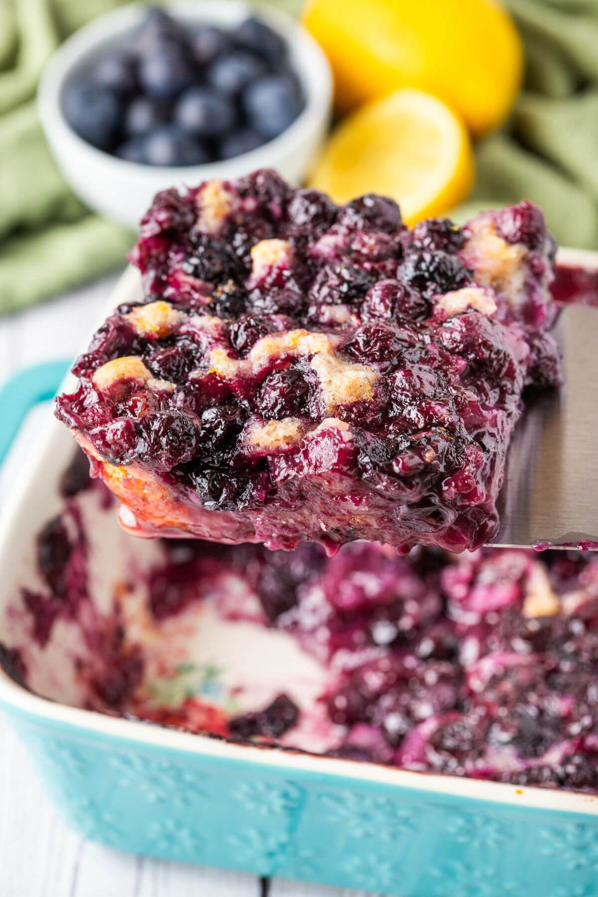 A piece of cobbler with berries is lifted into the air above a pan by a metal spatula.