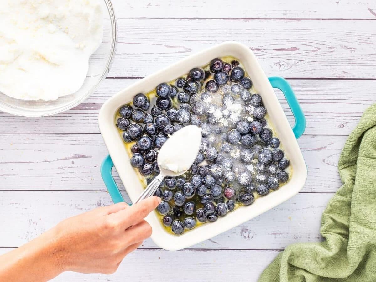 A hand uses a normal spoon to sprinkle sugar on top of a pan of blueberry dessert.