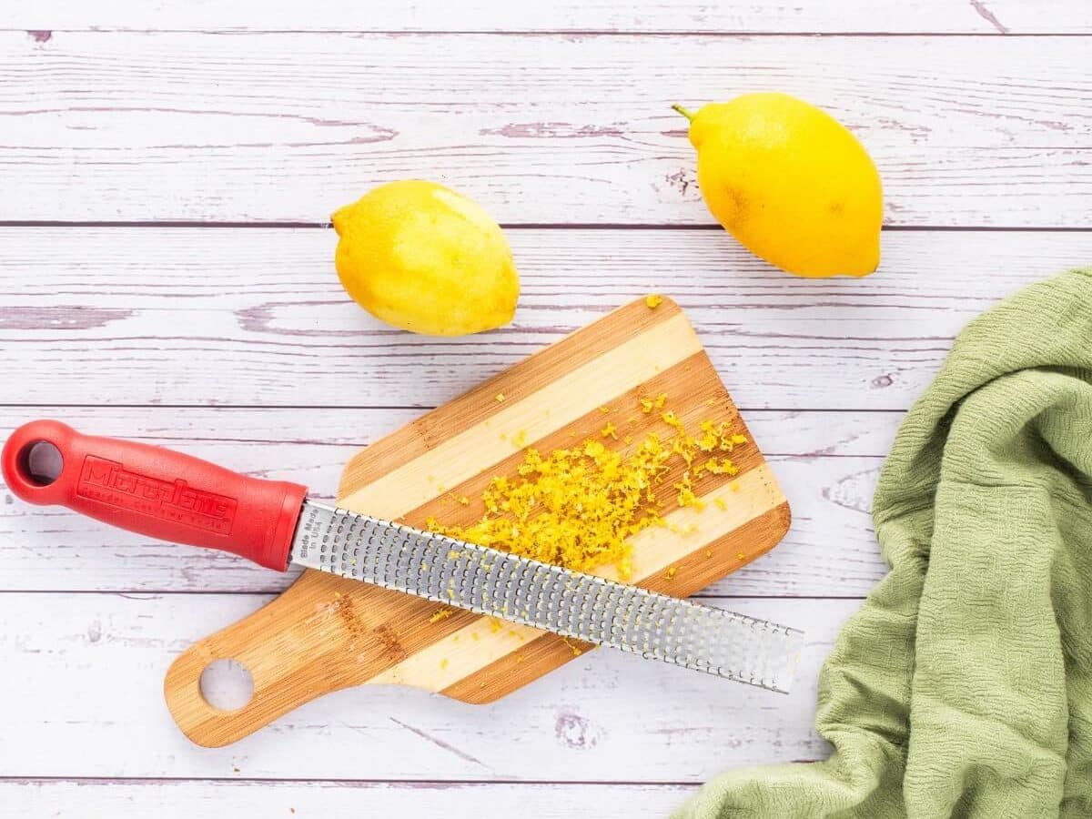 A metal zester sits on a wooden cut board with lemon zest resting on it.