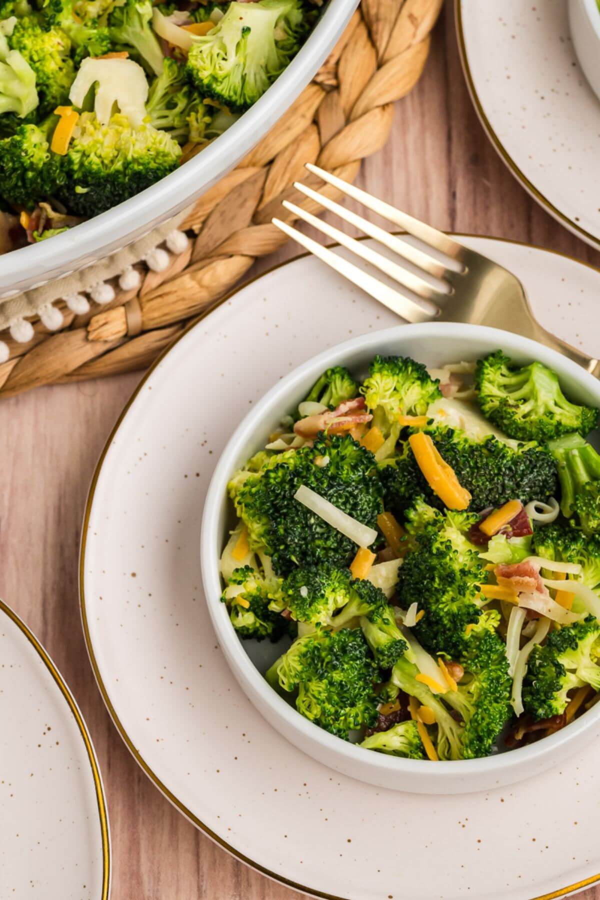 A white saucer frames a small white bowl filled with broccoli, cheese, and bacon next to a part view of the big bowl.