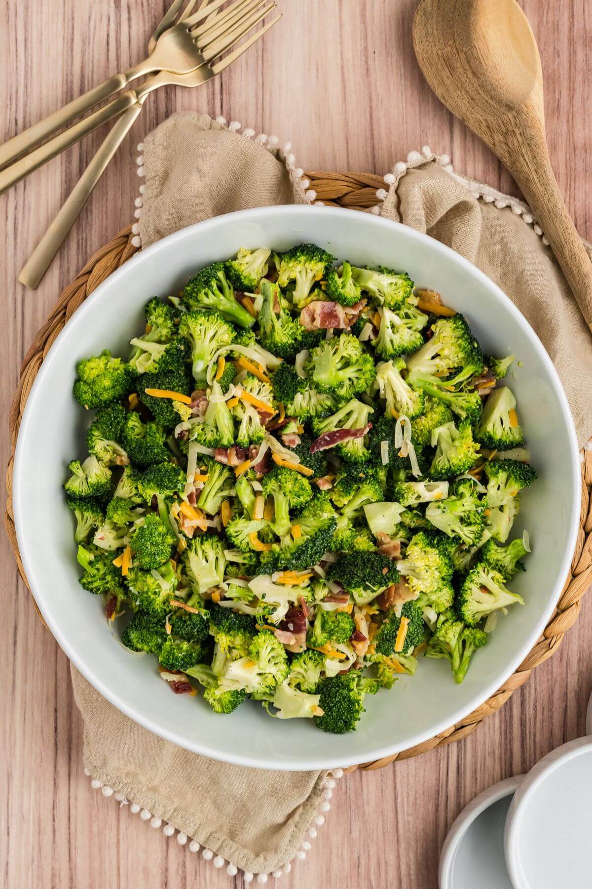 A large shallow serving bowl is filled with bright green broccoli florets, shredded cheese, and bacon chunks with utensils and a fine cloth.
