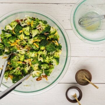 An empty glass bowl with a whisk and golden residue sits next to salt and pepper by a large bowl of broccoli, bacon, and cheese tossed with dressing.