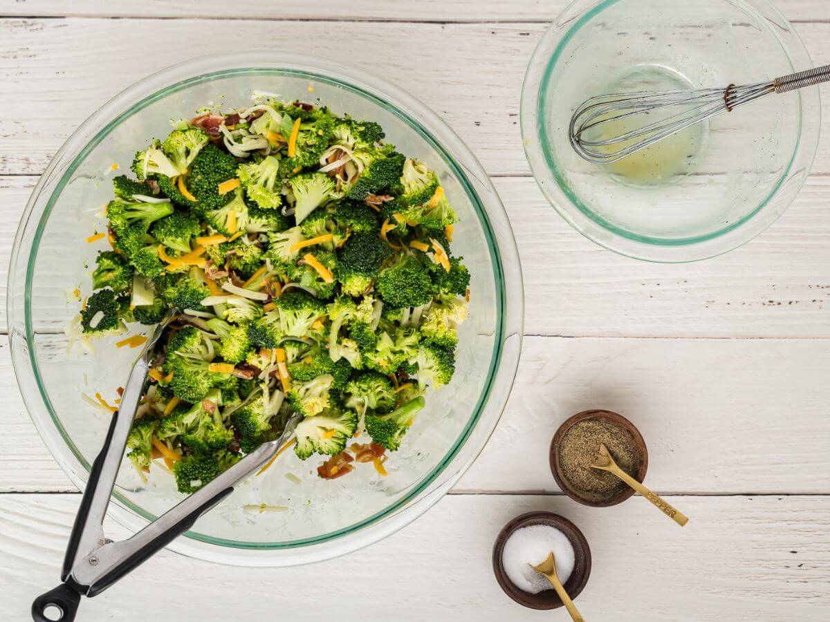 An empty glass bowl with a whisk and golden residue sits next to salt and pepper by a large bowl of broccoli, bacon, and cheese tossed with dressing.