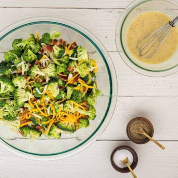 Grated white and yellow cheese sit on top of bright green broccoli and bacon pieces next to a bowl of mixed dressing.