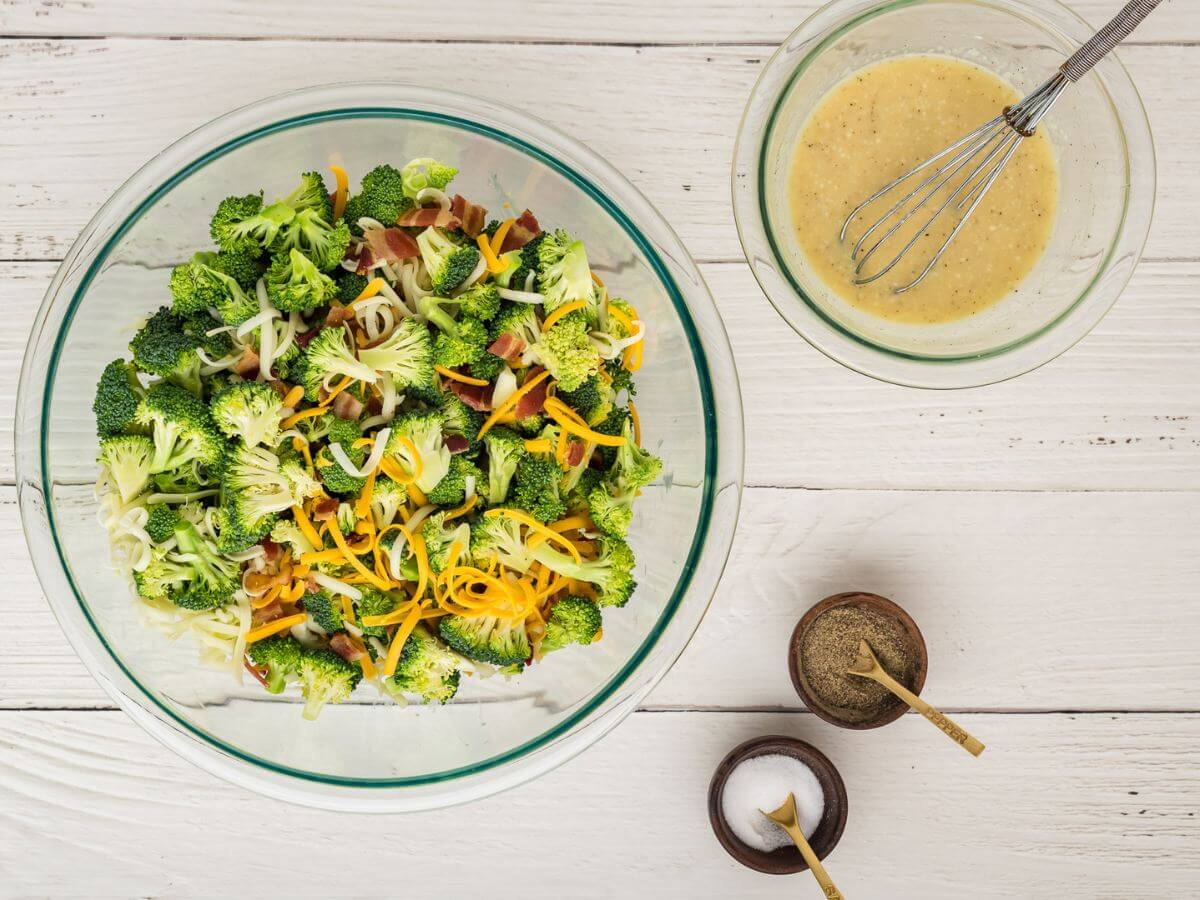 Grated white and yellow cheese sit on top of bright green broccoli and bacon pieces next to a bowl of mixed dressing.