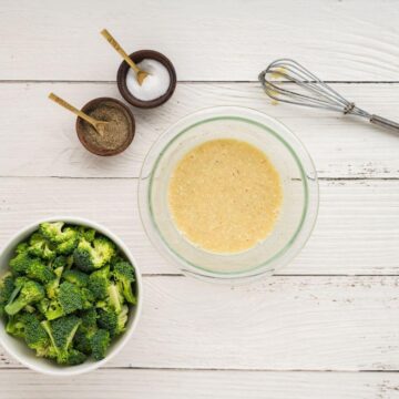 A whisk sits next to a glass bowl of dressing and by broccoli, salt, and pepper.