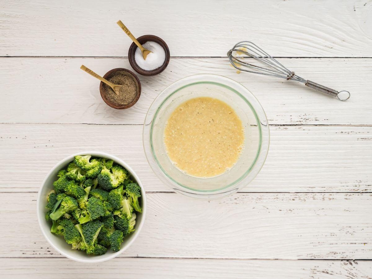 A whisk sits next to a glass bowl of dressing and by broccoli, salt, and pepper.