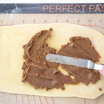 A hand is using a thin metal spatula to spread out the cinnamon filling on dough.