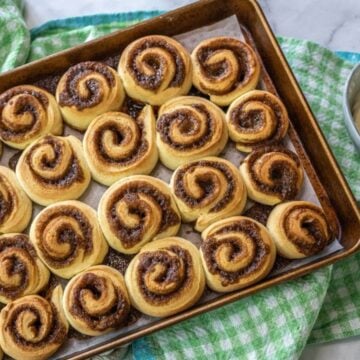 A baked pan of cinnamon buns sits on a checkered dishcloth by a bowl of icing.
