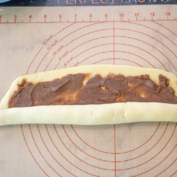 Cinnamon filling is inside flat dough that is being rolled up.