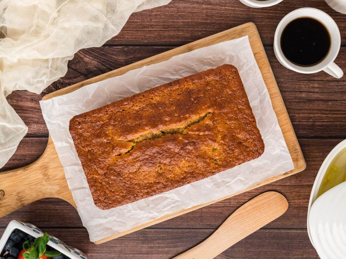 A loaf of banana bread rests on a lined cutting board next to coffee.