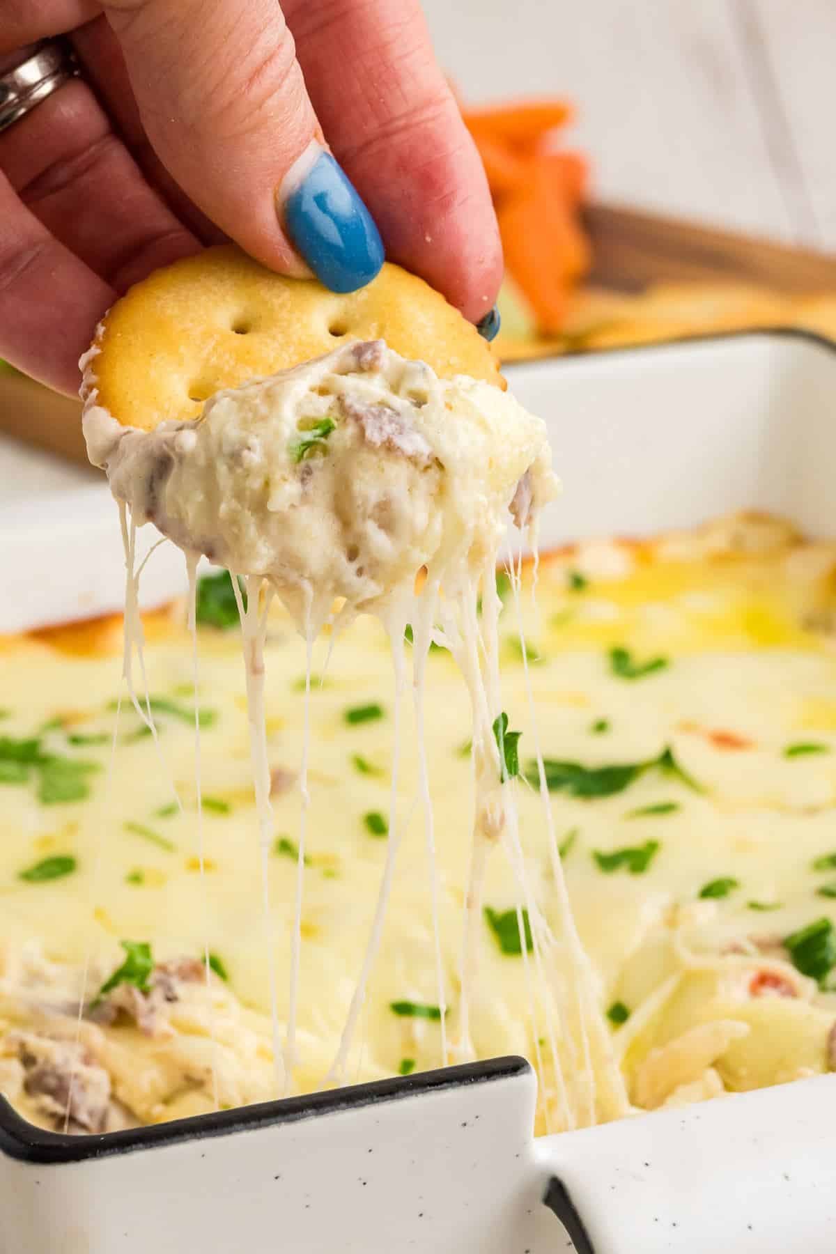 Hand dipping butter cracker into cheesy steak and cheese dip.