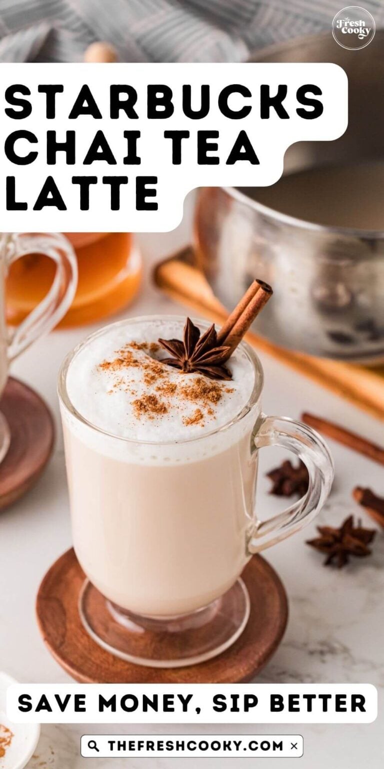 Looking down on a frothy topped mug of hot chai latte, topped with a sprinkle of cinmamon, a star anise and cinnamon stick, with a pot in the background, to pin.
