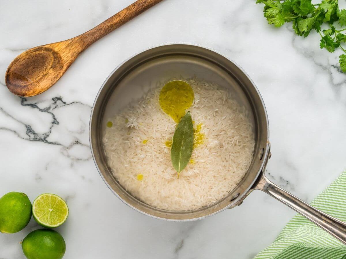 A saucepan is filled with water, oil, rice, and a bay leaf next to a wooden spoon and more garnish.