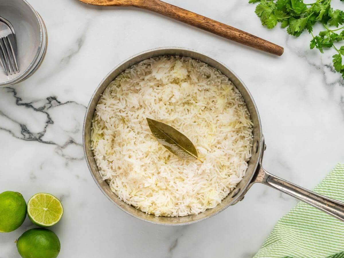 Fluffy cooked white rice sits in a saucepan with a bay leaf next to wooden spoon, fruit, and herbs.