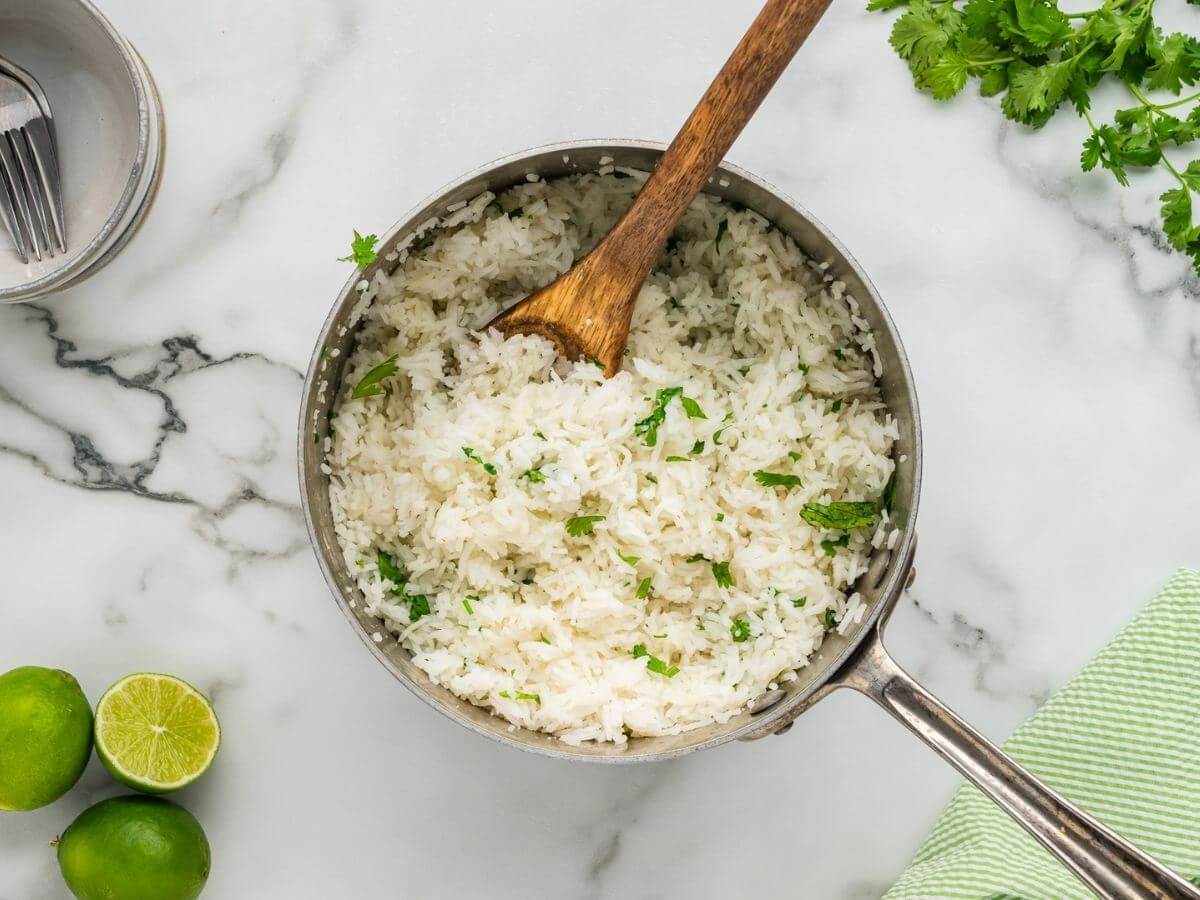White rice with green garnish sits in a metal saucepan with a wooden spoon inside.