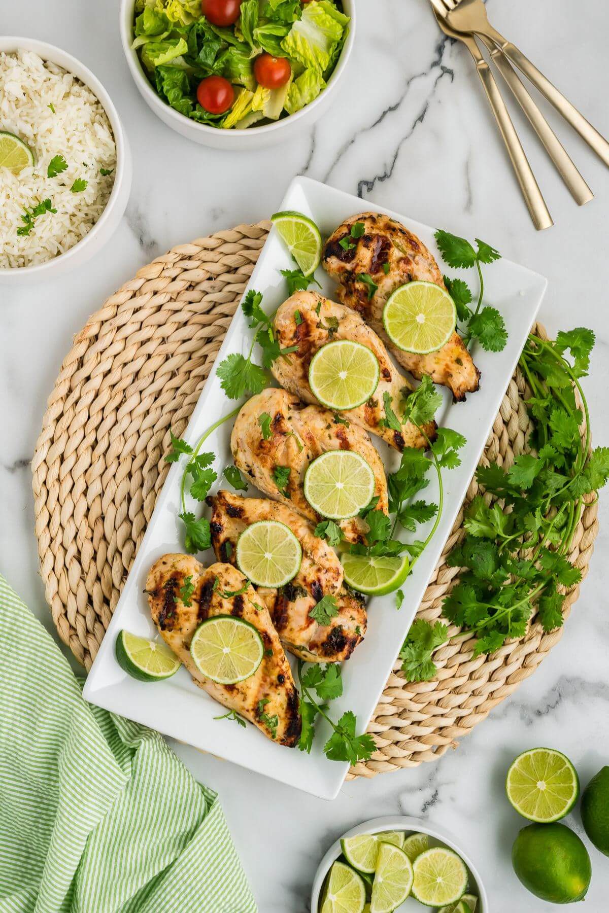 A round wicker placemat has a long serving plate of grilled, garnished chicken breasts next to salad and rice.